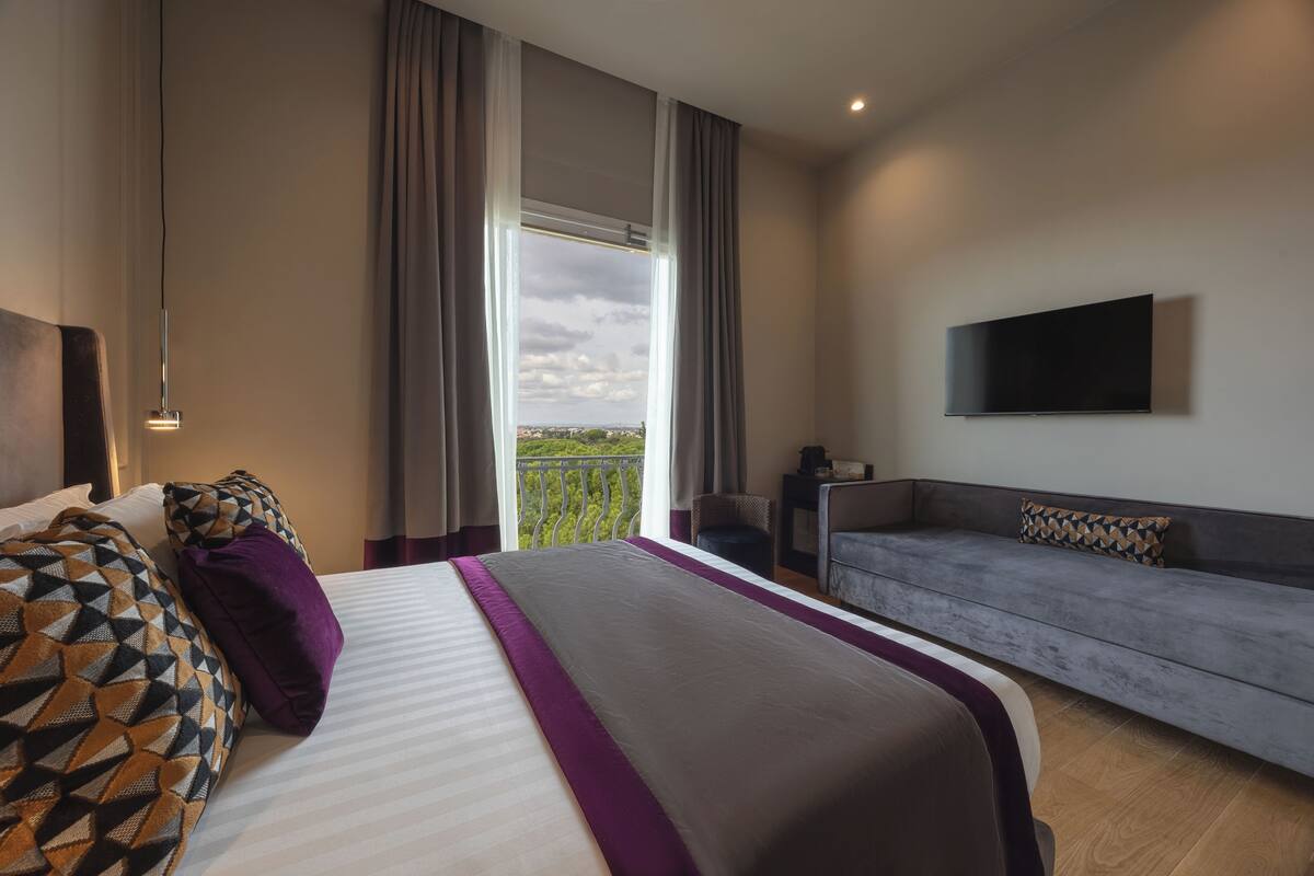 A hotel room with a large bed, a grey sofa, a TV mounted on the wall, and a balcony with a view.
