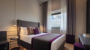 A modern hotel room with a large bed, a bedside table, a desk, and a view of the city. - Relais Pinciana (Rome)