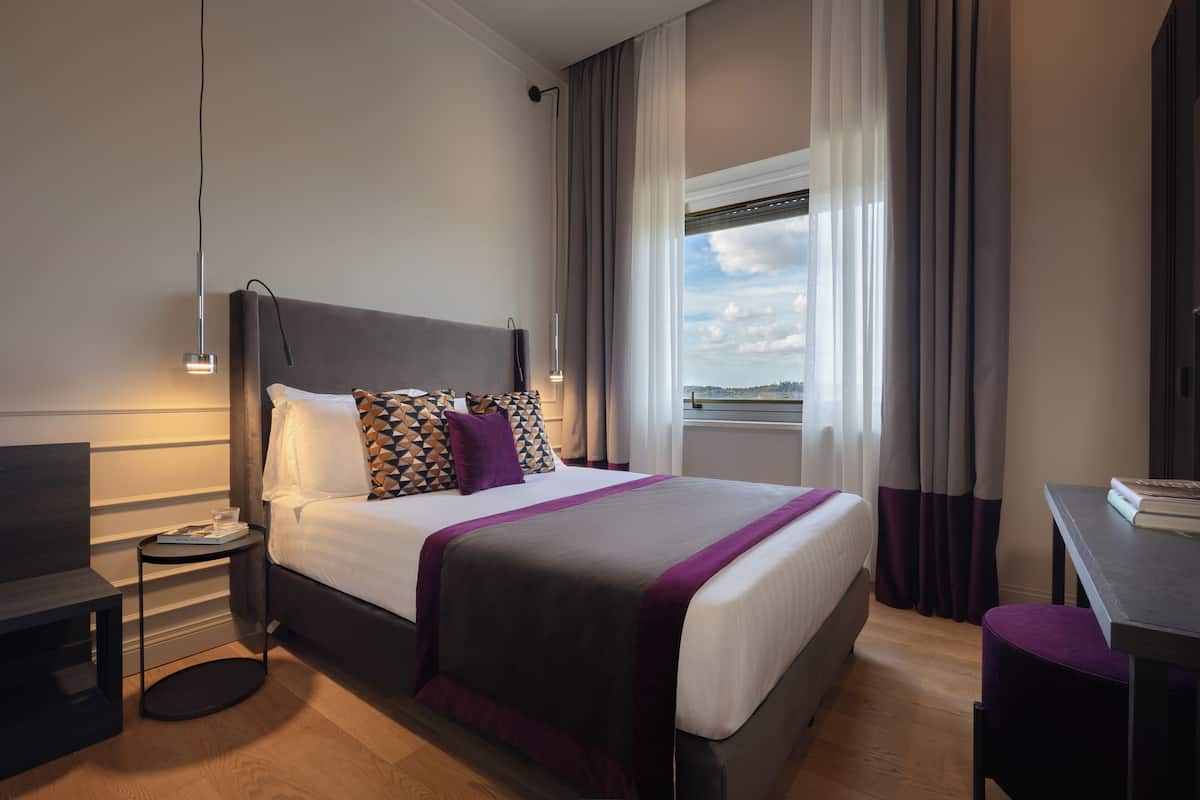 A modern hotel room with a large bed, a bedside table, a desk, and a view of the city.