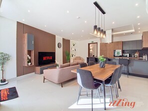 A modern living room with a flat-screen TV, a sofa, a dining table with chairs, and a kitchen area with appliances. - 🏡 Rooftop 3BR | Plunge Pool, BBQ & Private Garage (Ħal Għaxaq)