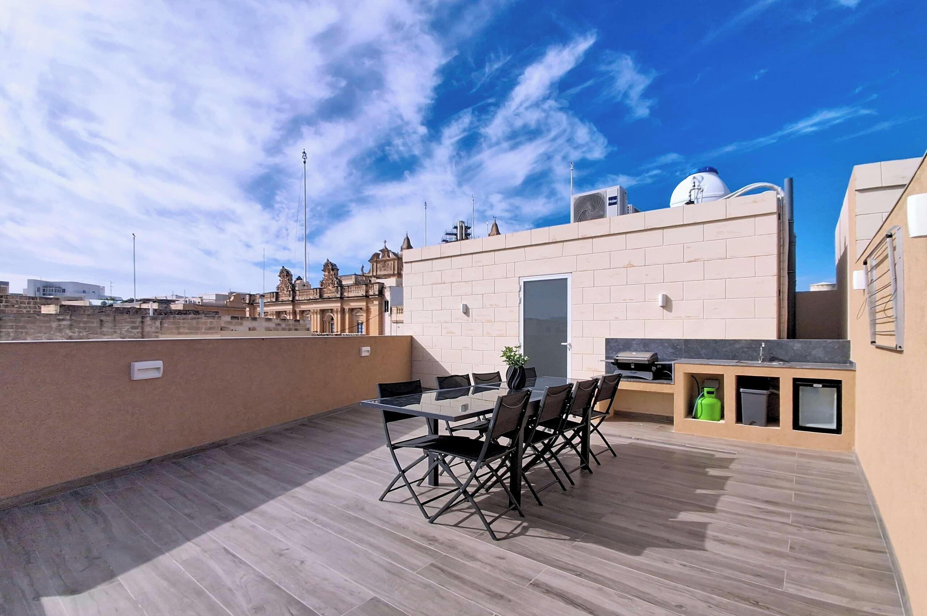 A rooftop terrace with a dining table and chairs, a small garden, and a view of a historic building.