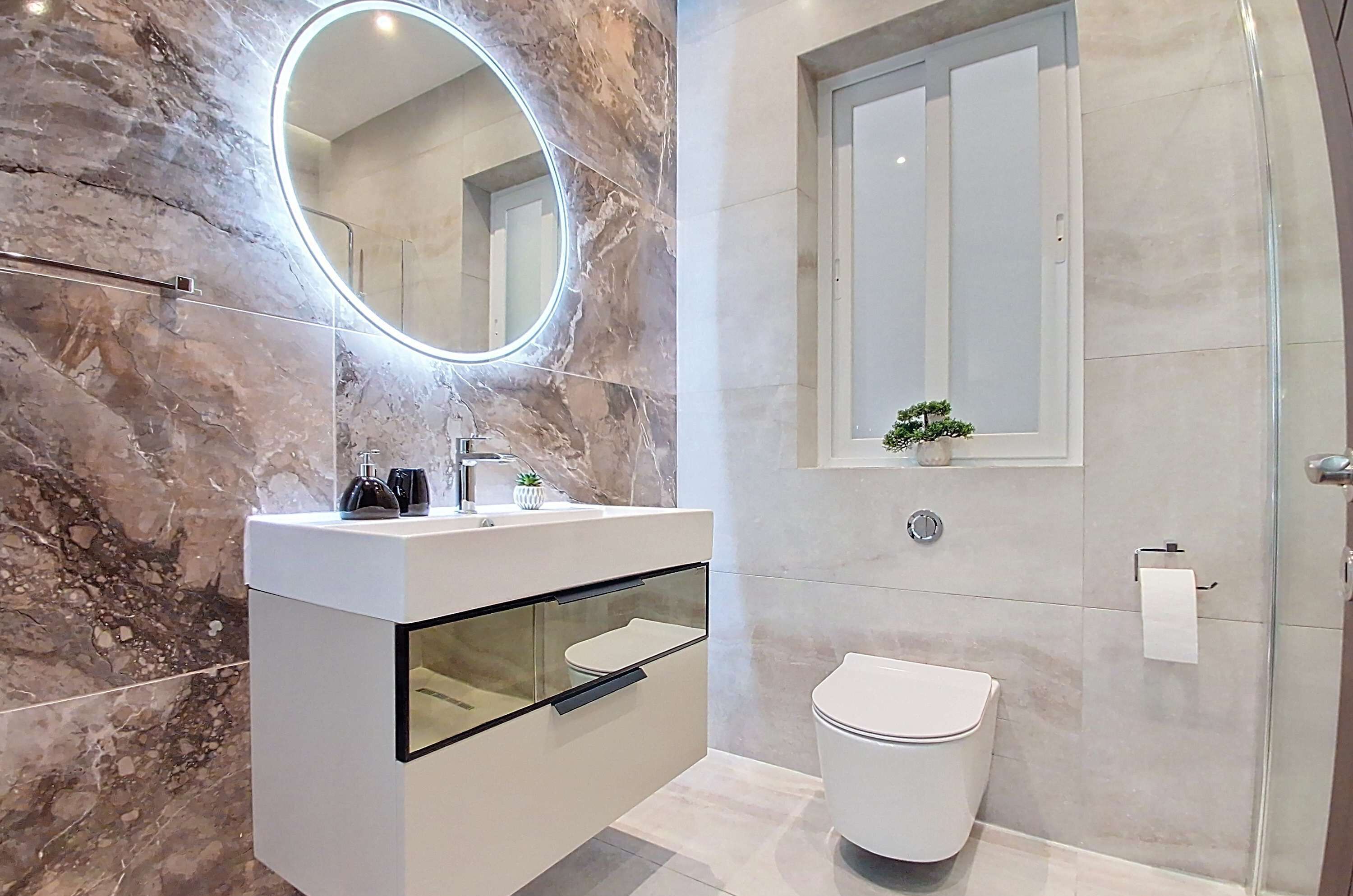 A modern bathroom with a marble wall, a round mirror with built-in lighting, a white sink with a glass enclosure, and a toilet.