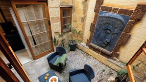 Une cour accueillante, avec des murs en pierre, une porte en bois, deux fauteuils en osier, une table où sont disposés des croissants et une grande sculpture en pierre.