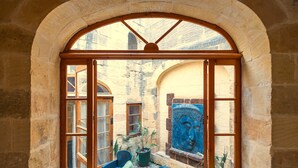 Une fenêtre en arc surmontée d’un cadre en bois, donnant sur une cour intérieure ornée d’une mosaïque bleue et de plantes en pot.