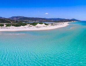 Un paysage côtier avec une plage de sable, des eaux turquoise et limpides, et des montagnes à l’horizon.