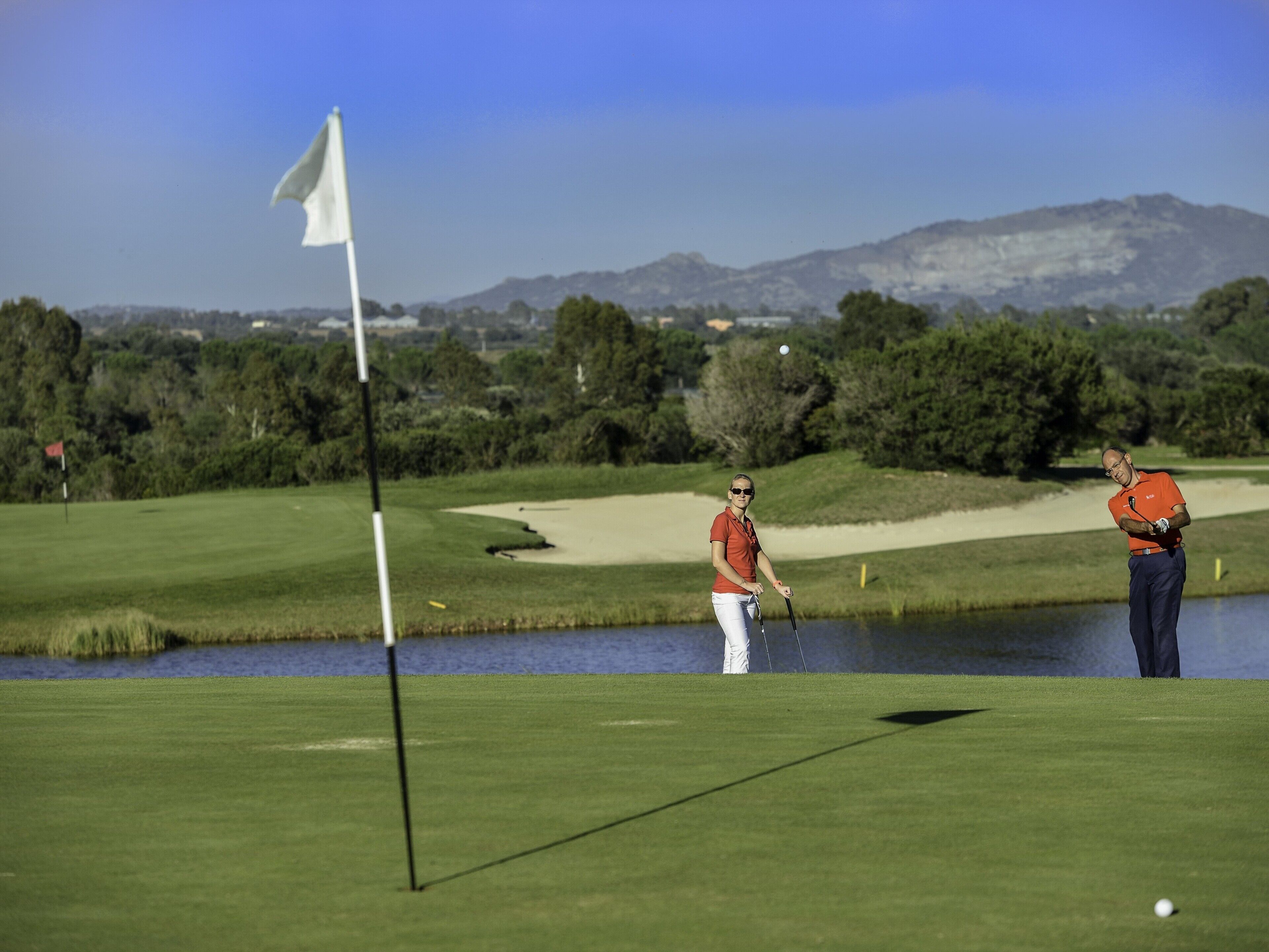 A golf course with a flag, water body, and distant mountains.