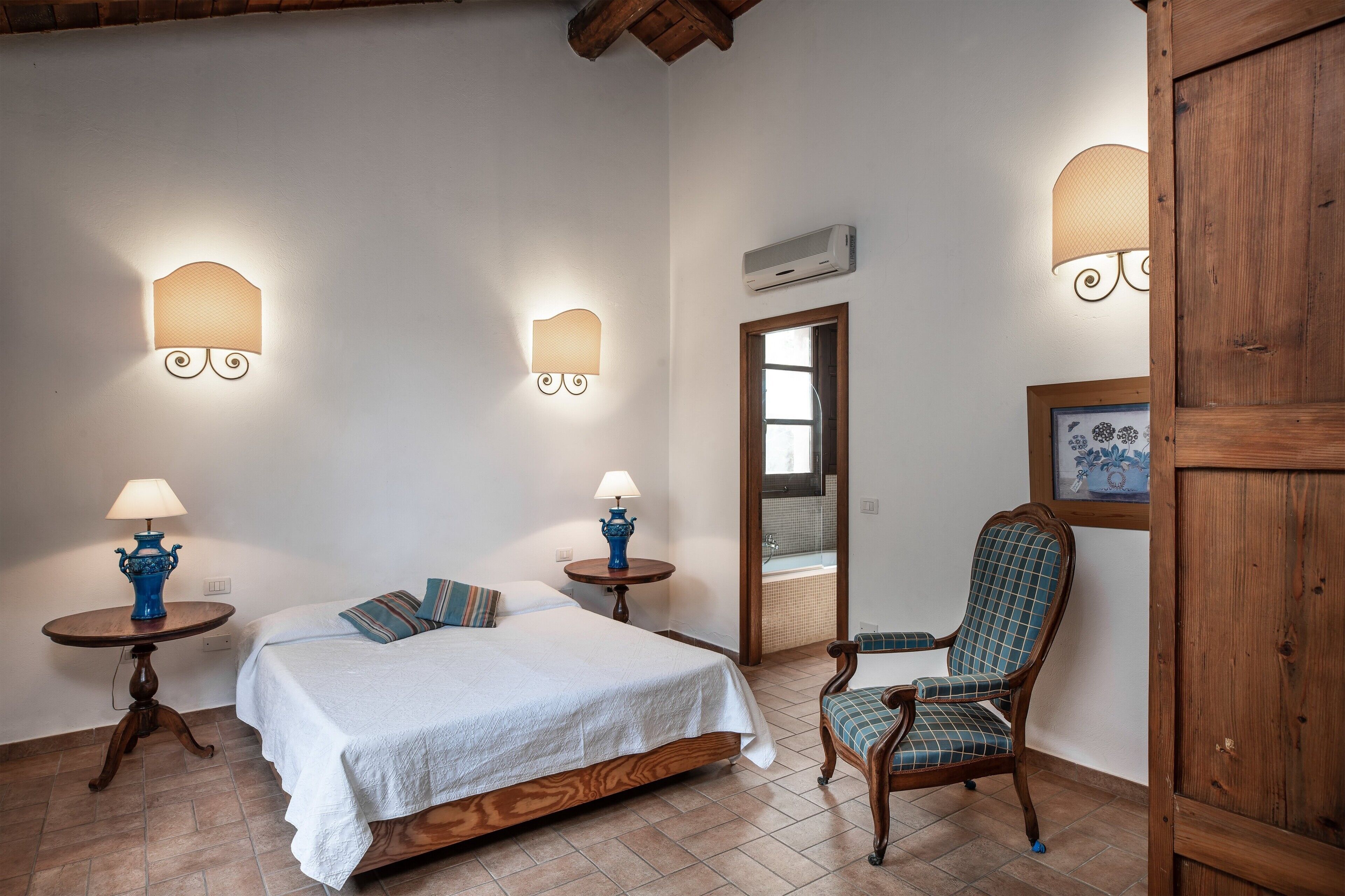 Een slaapkamer met een bed, twee nachtkastjes met lampen, een stoel, een houten garderobe en een badkamer die door een deuropening zichtbaar is.