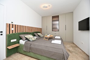 Une chambre moderne avec un lit recouvert d’un tissu vert, une tête de lit en bois, une table de chevet, un plateau avec des articles pour le petit-déjeuner et une armoire avec un miroir.