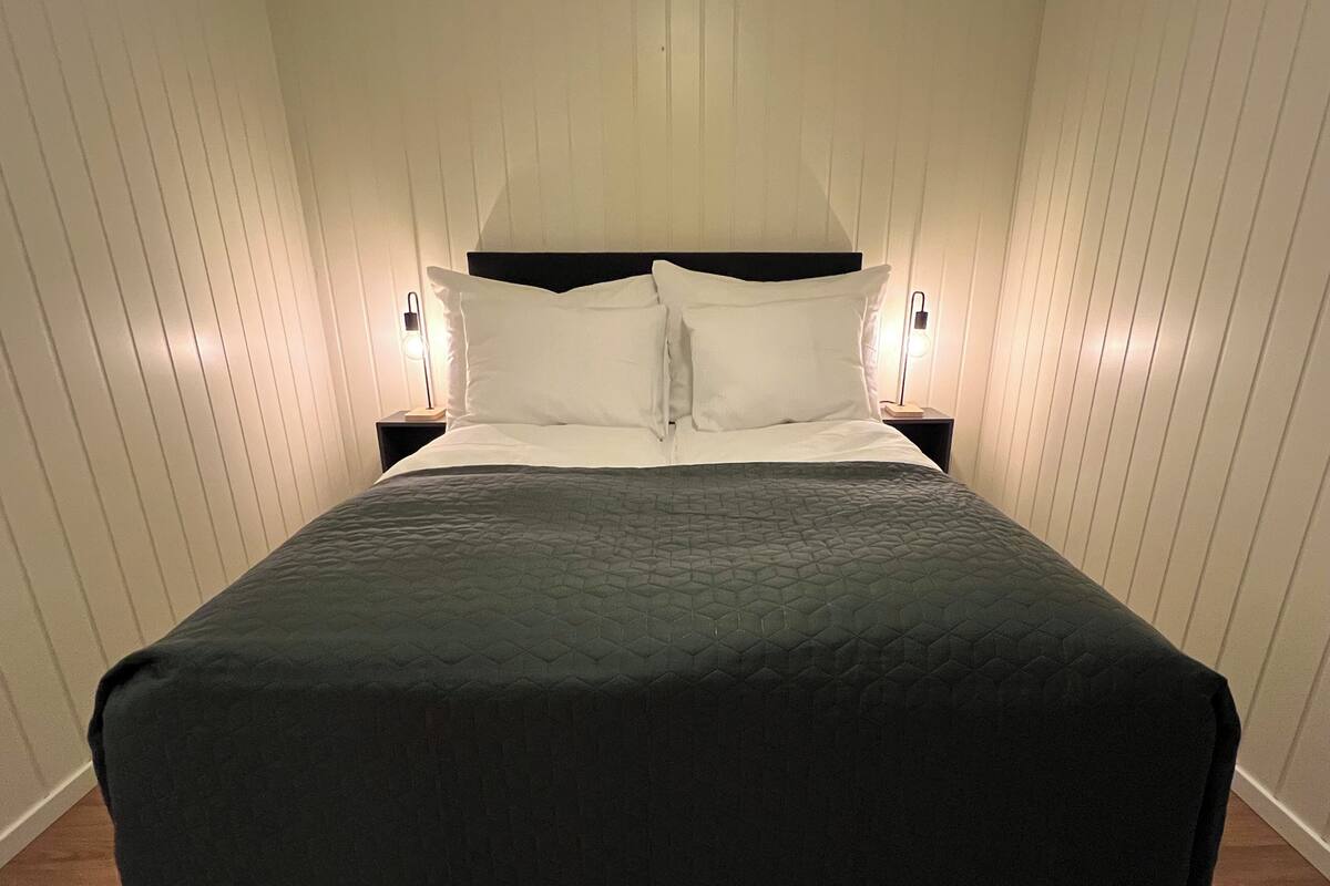 Una cama bien hecha con un edredón oscuro, dos lámparas de noche y un cabecero de madera.