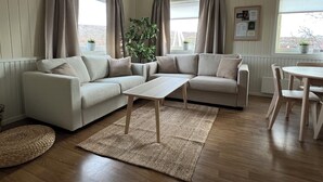 A living room with a beige sofa, a coffee table, and a dining table with chairs. There are two windows with curtains, a potted plant, and a framed picture on the wall. - Tanabrew  (Tana)