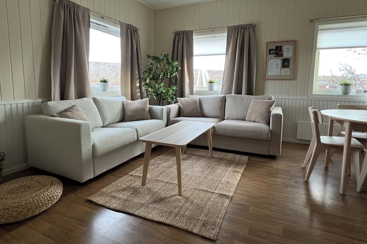 Una sala de estar con un sofá color beige, una mesa de centro y una mesa de comedor con sillas. Hay dos ventanas con cortinas, una planta en maceta y un cuadro en la pared.