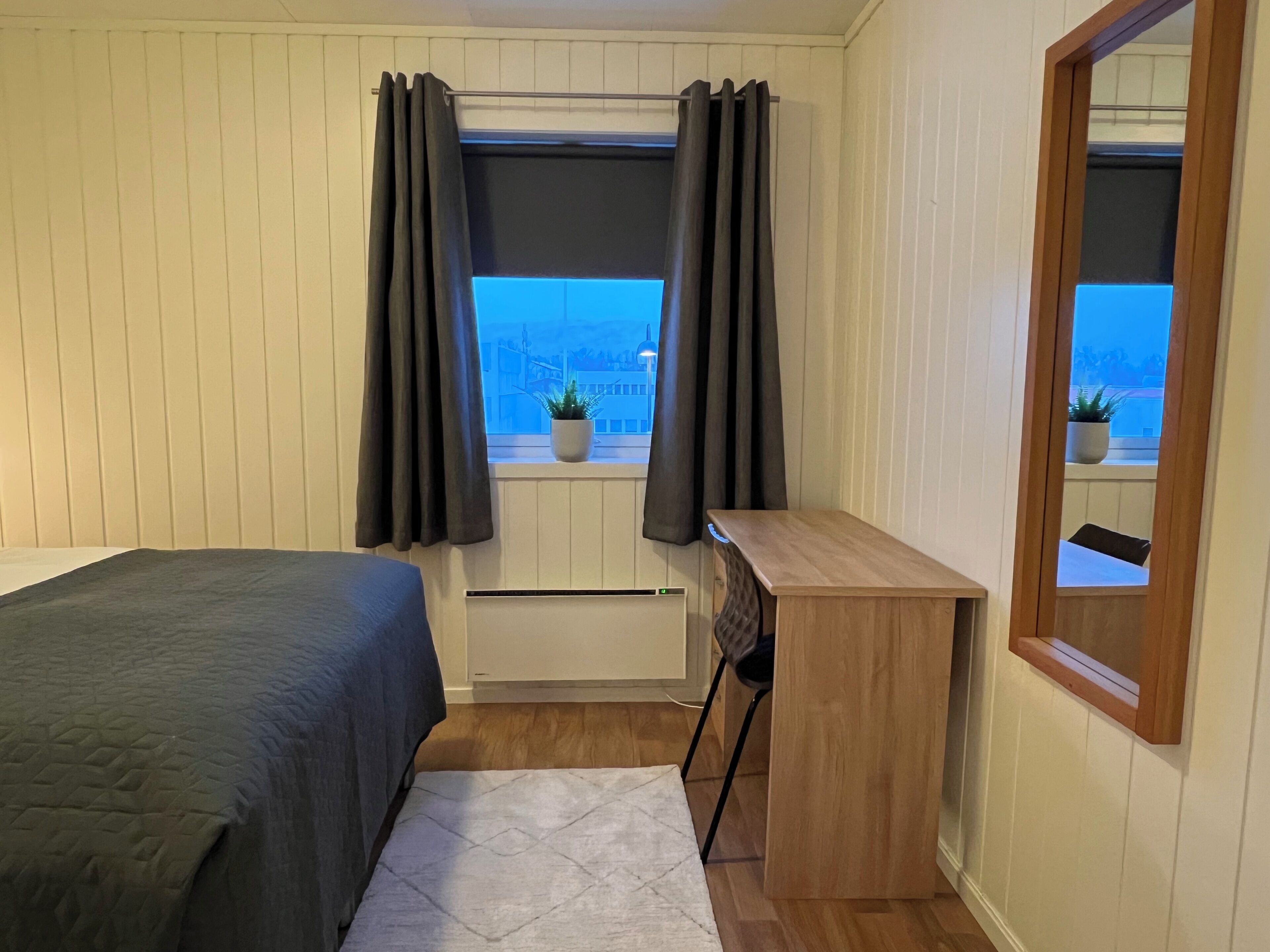A bedroom with a bed, a desk, a chair, a window with curtains, a radiator, and a mirror.