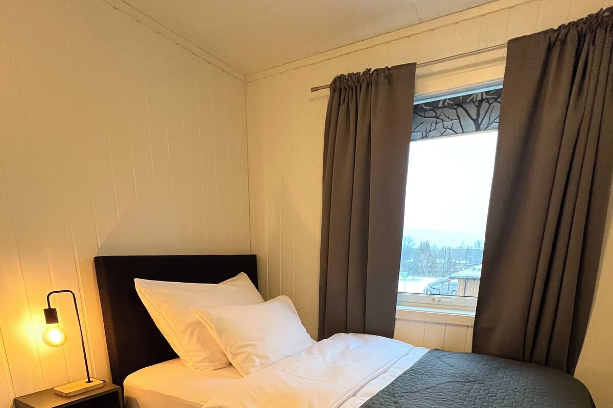 Una cama bien hecha con sábanas blancas, un cabecero oscuro y una colcha acolchada. Hay una mesita de noche con una lámpara y un pequeño estante. Una alfombra color beige cubre el suelo, y la habitación tiene una ventana con cortinas.