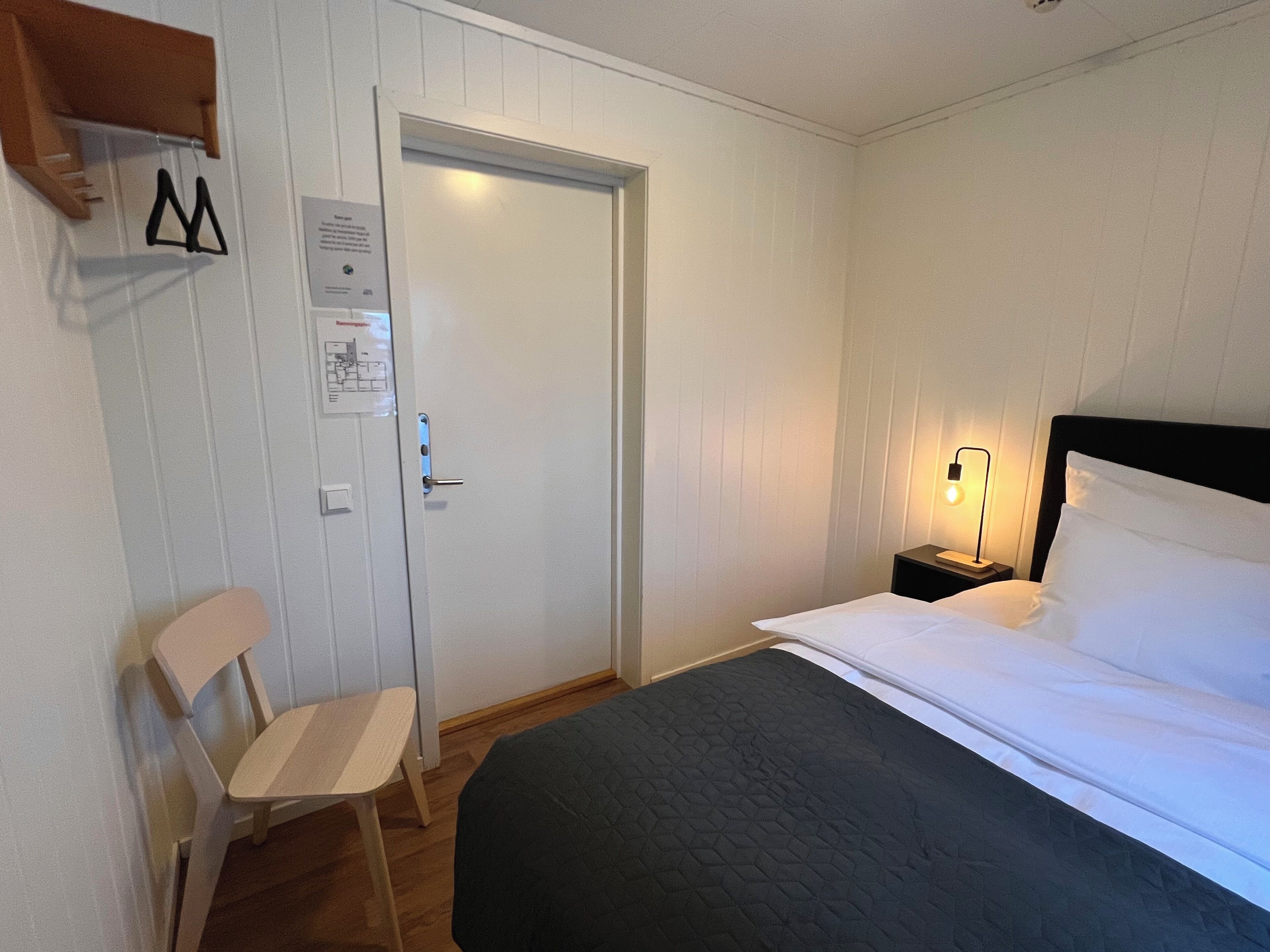 A bedroom with a bed, a chair, a nightstand with a lamp, and a door with a sign.