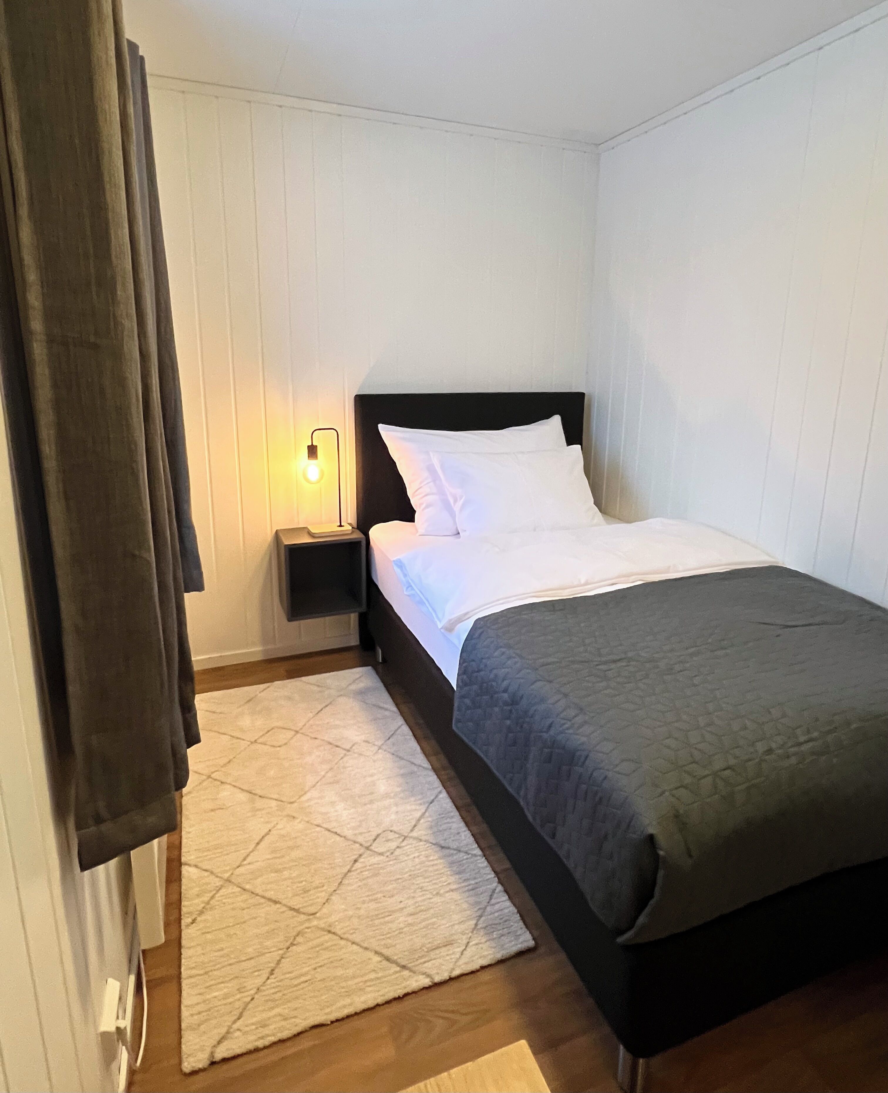 A neatly made bed with white linens and a dark headboard, a bedside lamp, and a patterned rug on a wooden floor.