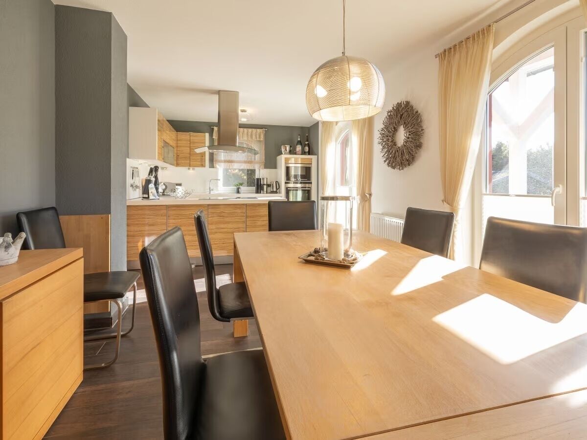 A modern dining area with a wooden table and black chairs, a kitchen in the background, and a large window with curtains.