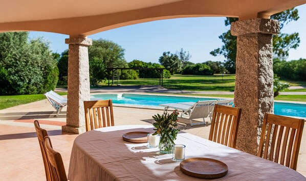 Un área de comedor al aire libre techada, con vista a una piscina, sillas de madera y una mesa lista para dos.