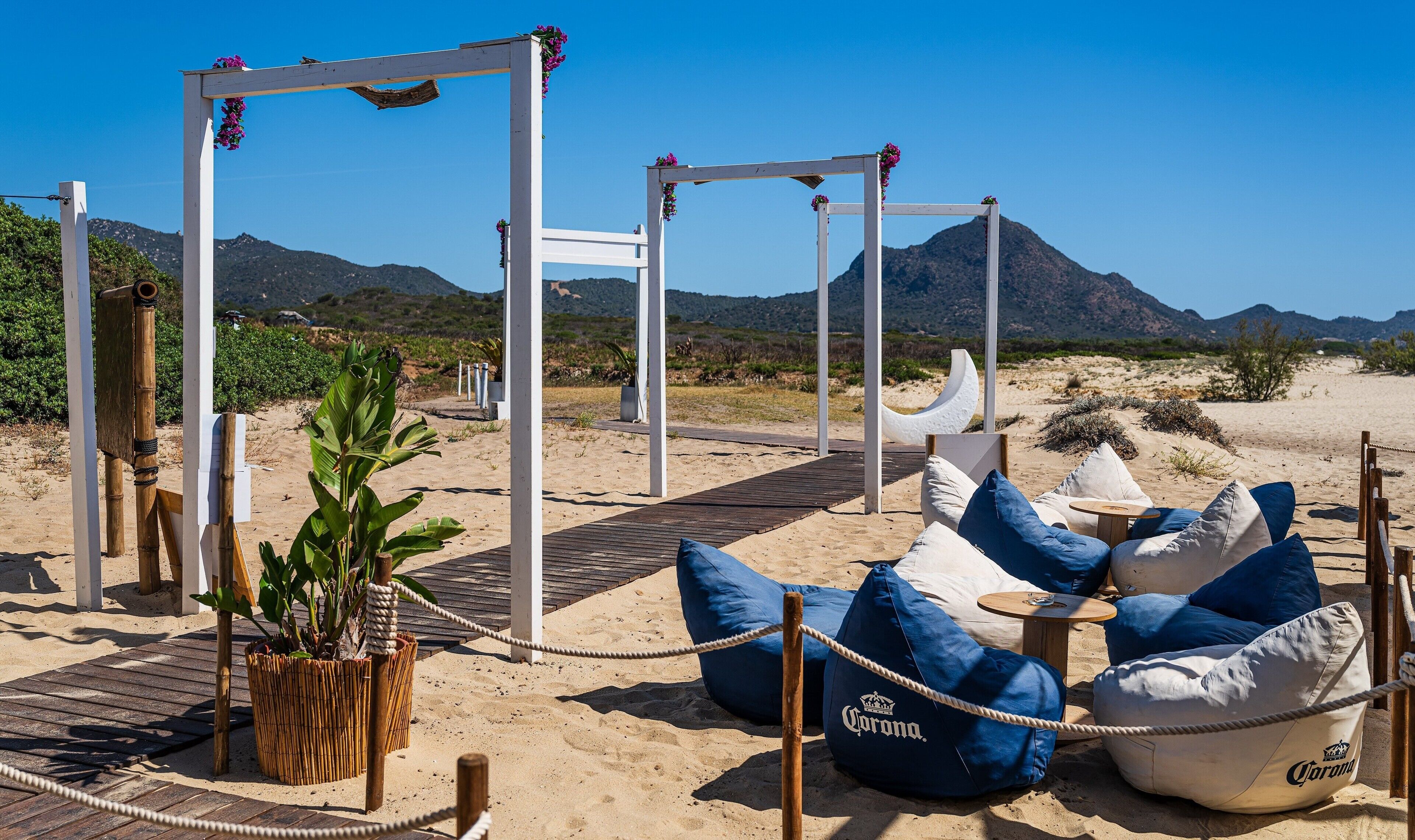 A beach setting with a wooden deck, white pergola, and bean bags labeled 'Corona.'