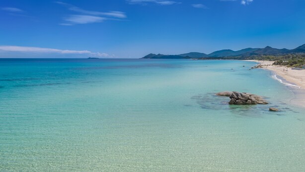 Ein Strand mit klarem türkisblauem Wasser, einem Felsen und Bergen im Hintergrund.