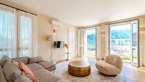 Una sala moderna con sofá, reposapiés y sillón, un televisor de pantalla plana y un amplio ventanal corredero que da a un balcón con vistas a la montaña.