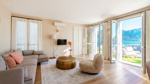Una sala moderna con un sofá, un reposapiés redondo, una silla y un amplio ventanal corredero que da a un balcón con vistas al mar.