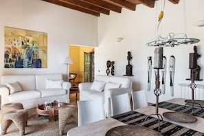 Un salon avec un canapé blanc, une table basse et une table à manger avec des chaises. Il y a un tableau au mur et un oiseau perché sur un luminaire.