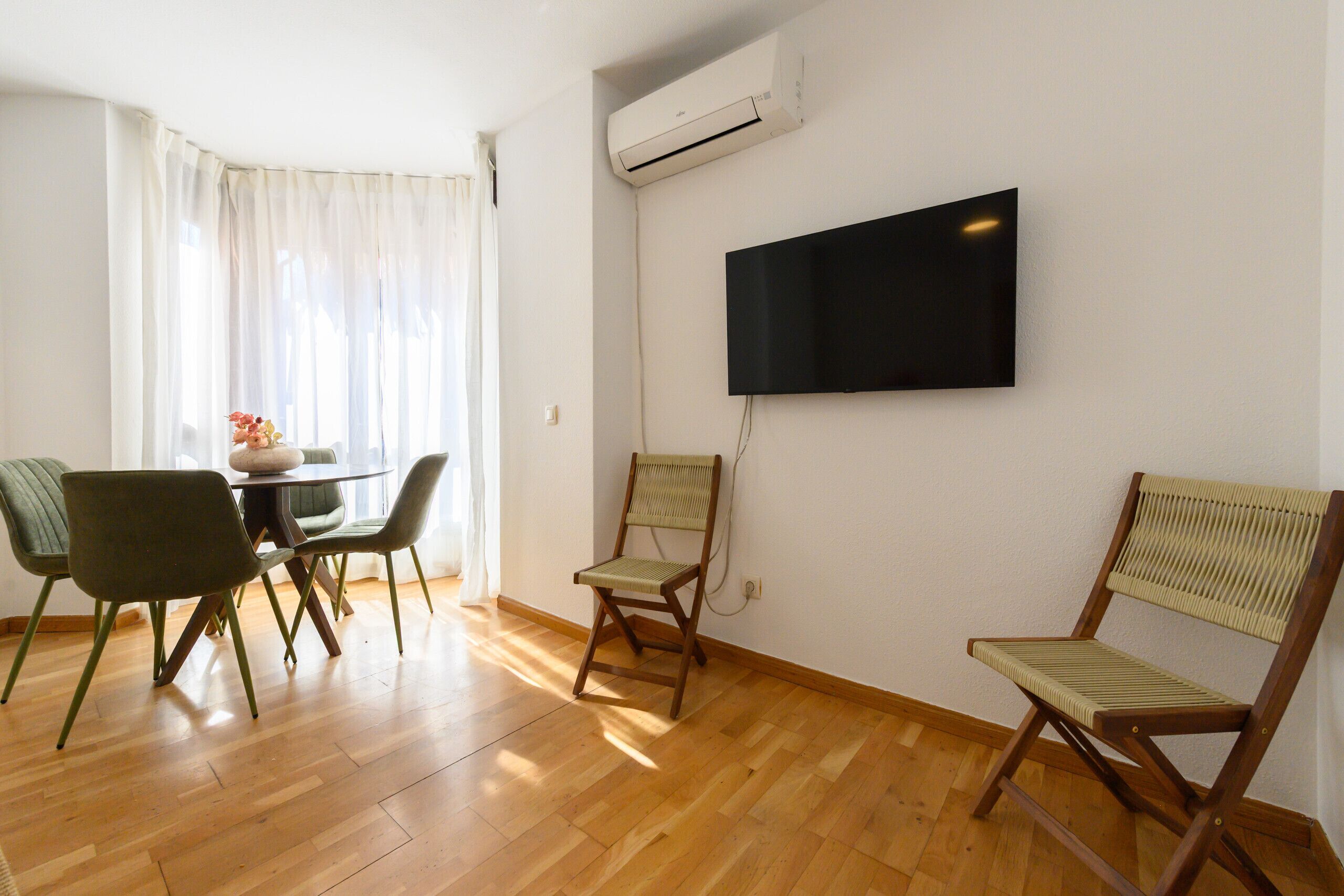 A room with a dining table and chairs, a flat-screen TV mounted on the wall, and an air conditioning unit.