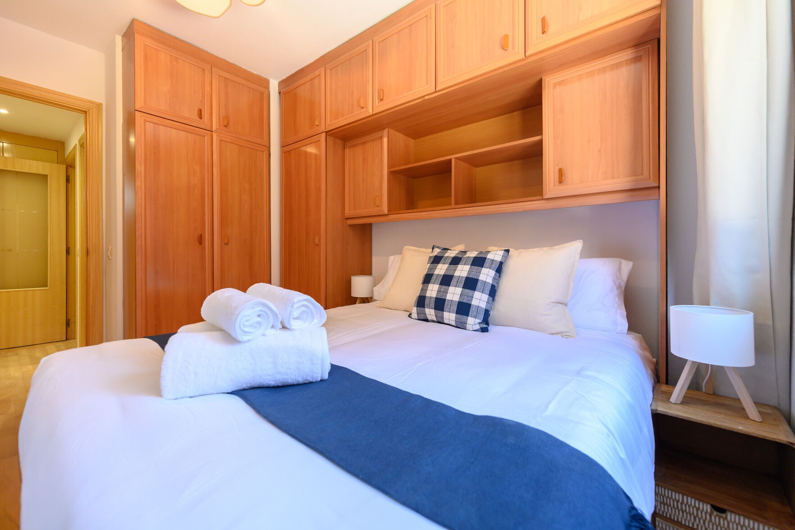 A neatly made bed with white linens and a blue throw blanket, wooden headboard, and a bedside lamp.