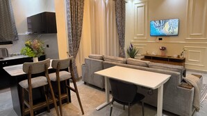 Una sala moderna con un sofá gris, una mesa de comedor con sillas, un televisor montado en la pared y una zona de cocina con microondas y una planta.