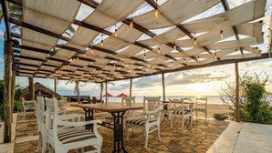 An outdoor seating area with stone flooring, white chairs, and a wooden table under a pergola with hanging lights. - Dolphin Beach Resort (Etalai)