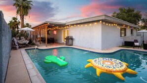 A swimming pool with inflatable toys, a covered patio with string lights, and a house with a visible garage. - Sleeps 16! Luxury Phoenix Retreat Near Biltmore- Pool, Theater & Game Room. (Phoenix)
