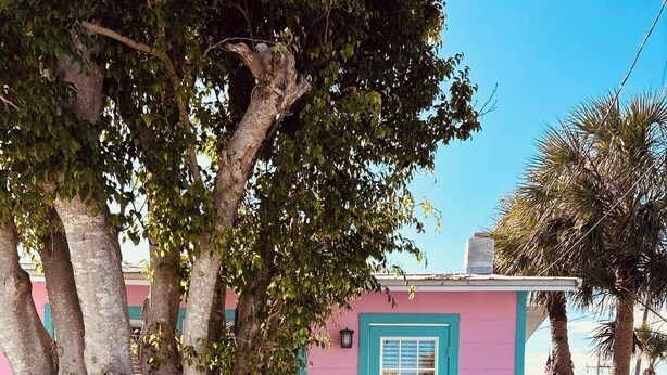 Une petite maison rose avec une porte et une fenêtre turquoise, entourée d’arbres et bordée d’une allée de gravier.