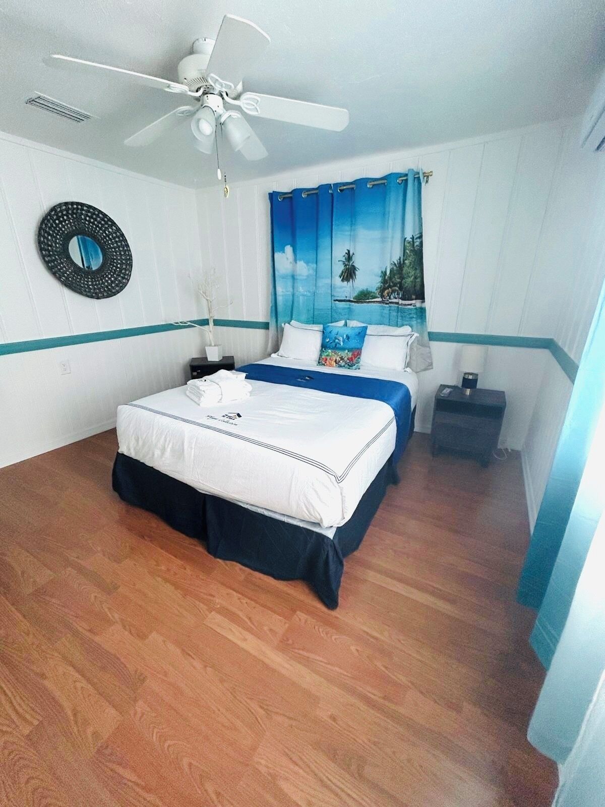Quarto com cama, ventoinha no teto, mesa de cabeceira e janela com vista para palmeiras.