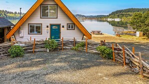 Une cabane triangulaire en bois avec une porte bleue, située au bord d’un lac, avec un quai et une table de pique-nique.