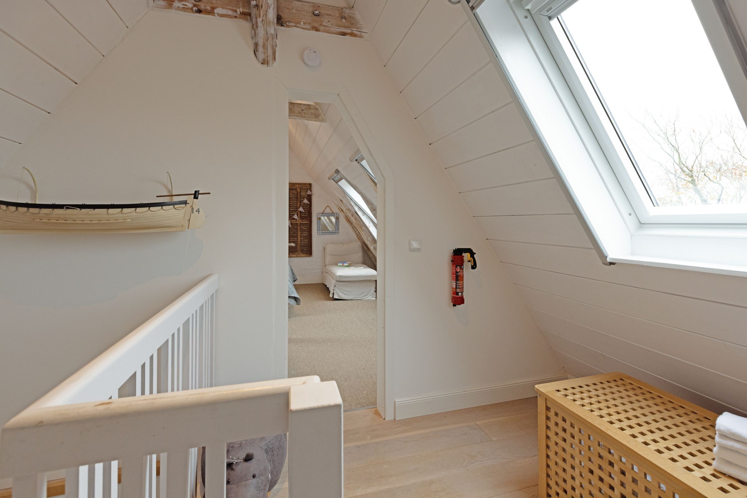 A room with a sloped ceiling, a skylight, a wooden crib, a laundry basket, and a small bed in the background.