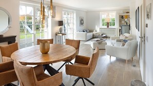 A spacious living area with a round wooden dining table surrounded by chairs, a sofa, and a coffee table. There is a large window providing natural light and a view of the outdoors. - Spacious vacation home Lüttje Butt with garden and terrace near the beach on Sylt (List auf Sylt)
