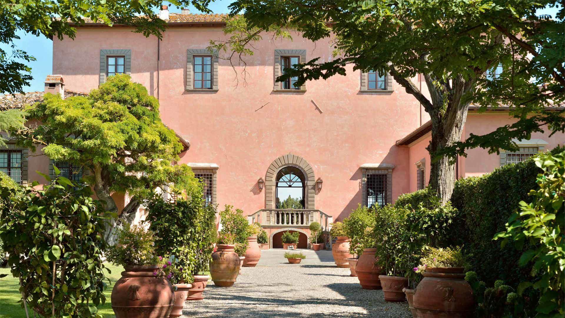 Une villa rose, dotée d’une arche centrale, et entourée de plantes en pot et d’arbres.