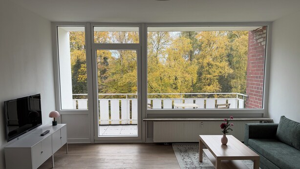 Sala de estar com televisão, mesa de centro, sofá e vista para árvores através de grandes janelas.