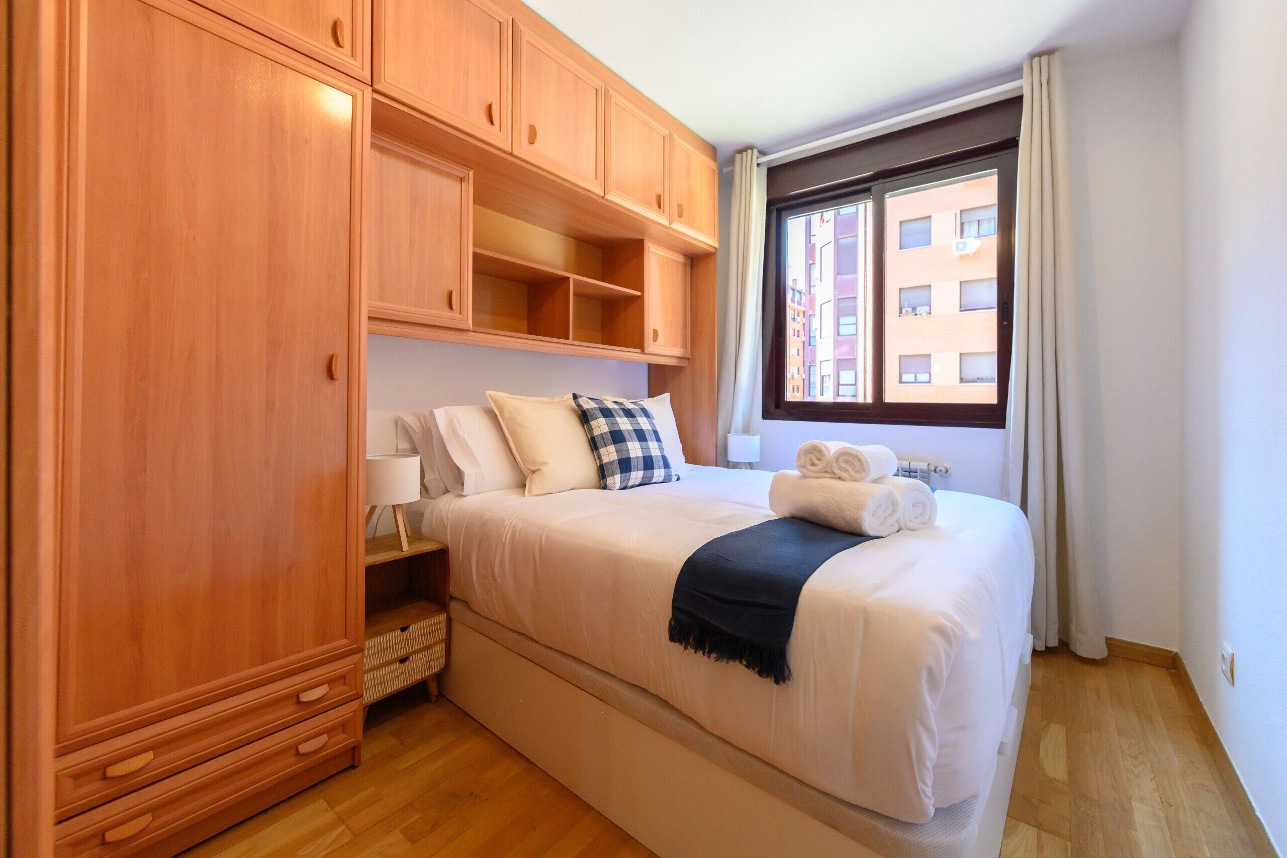 Une chambre avec un grand lit, une armoire en bois et une fenêtre donnant sur des immeubles.