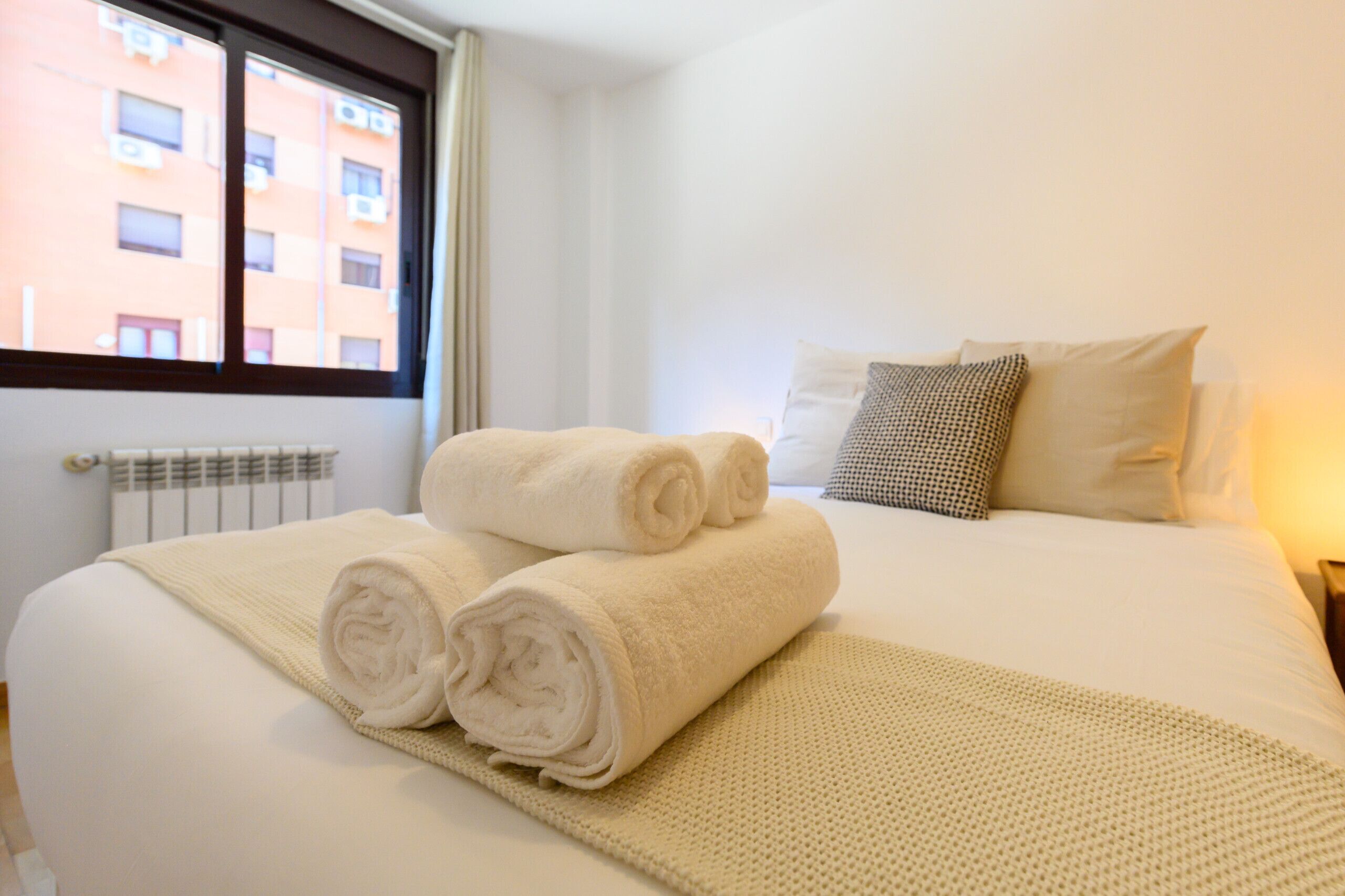A neatly made bed with rolled white towels, a beige throw blanket, and two pillows.