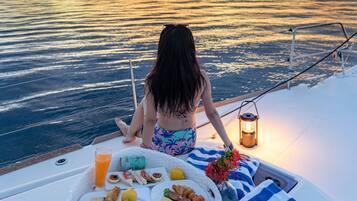 A person on a boat with a table of food and drinks, a lantern, and a view of the water.