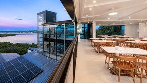Un restaurant moderne doté d’une terrasse donnant sur un plan d’eau, avec des panneaux solaires, des tables en bois et une façade en verre.