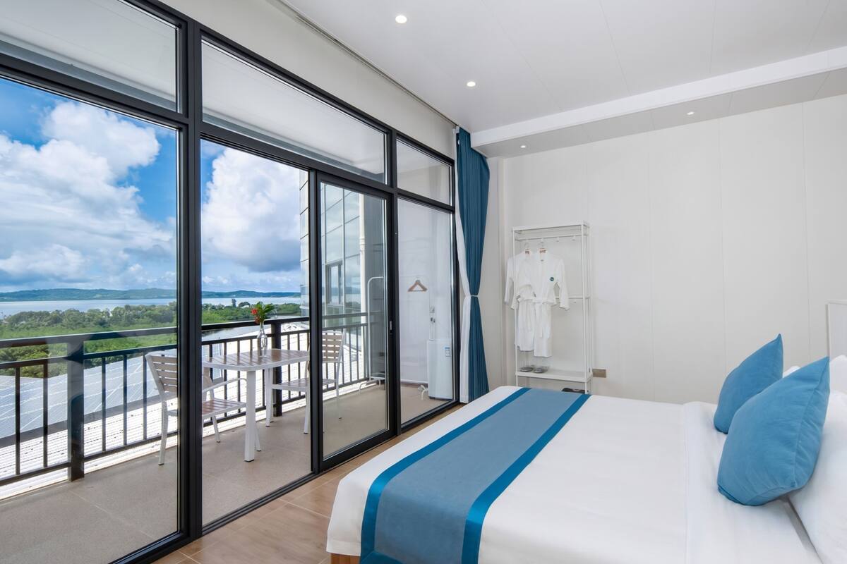 Ein Hotelzimmer mit großem Balkon, einem Bett mit blauer Bettwäsche und Blick auf ein Gewässer.