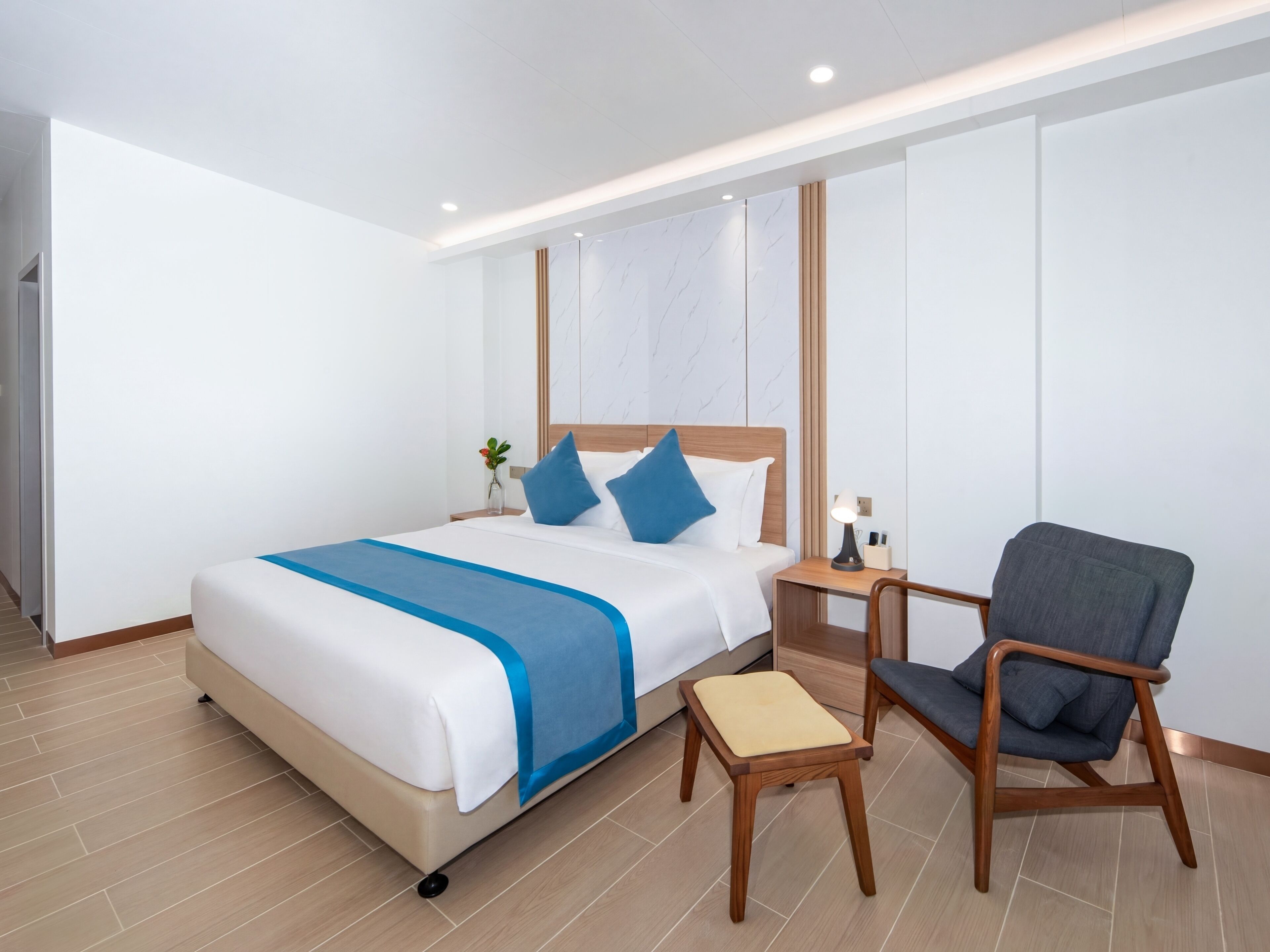 A modern hotel room with a bed, bedside table, chair, and a small table with a lamp.