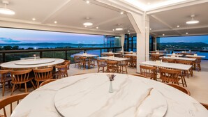 Un restaurant avec des tables rondes en marbre et des chaises en bois, de grandes fenêtres donnant sur un plan d’eau, et un plafond haut avec des luminaires encastrés.