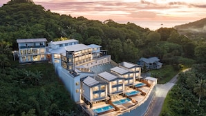Une maison moderne à plusieurs étages, dotée d’une piscine, entourée d’une végétation luxuriante et de collines.