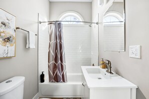 A bathroom with a white toilet, a bathtub with a shower curtain, a sink with a faucet, and a mirror above the sink. - Sleek Modern Indy Home w/ Yard, BBQ & Easy Access (Indianapolis)