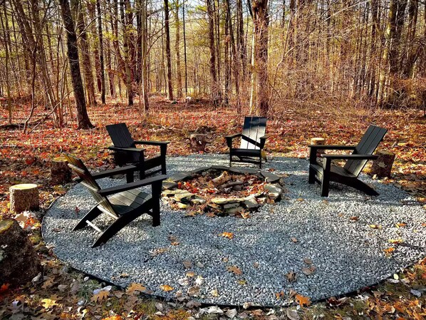 A circular seating area with three Adirondack chairs around a central fire pit, set on a gravel surface in a forested area.