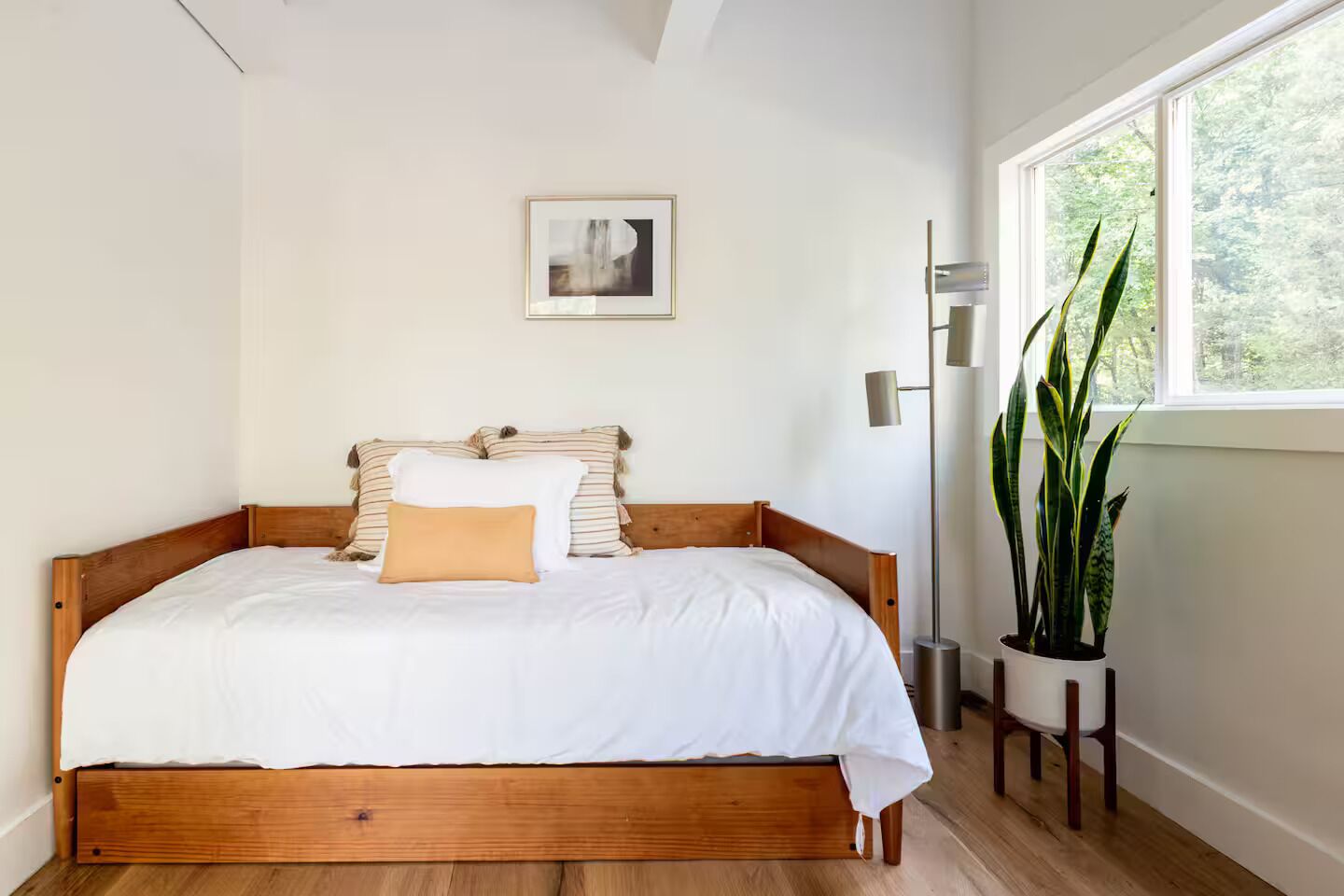 A bedroom with a wooden bed, white bedding, a framed picture on the wall, a floor lamp, and a potted plant by the window.