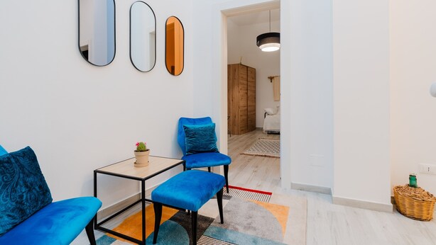 Una sala de estar moderna con un sofá, una mesita y un sillón azul. Hay tres espejos en la pared, una lámpara colgante y un aparador de madera al fondo.